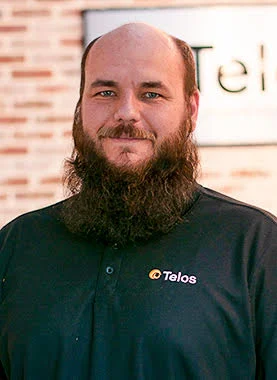 A man with a full beard and a receding hairline, wearing a black Telos shirt, stands in front of a blurred brick wall and a sign.