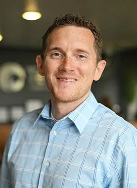A man with short brown hair wearing a light blue plaid shirt smiles while standing indoors in a softly lit setting.