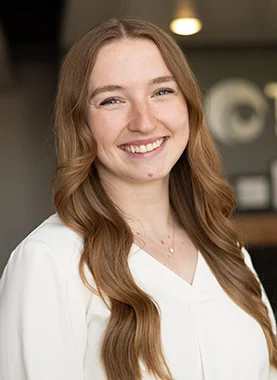 A woman with long, wavy light brown hair is smiling, wearing a white blouse, standing indoors with a blurred background.