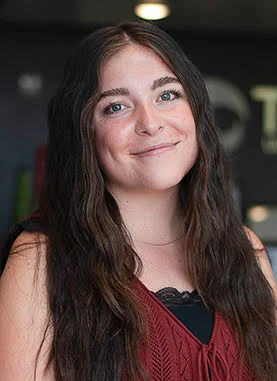 Woman with long wavy brown hair and blue eyes smiling, wearing a maroon sleeveless top with a black lace underlayer, standing indoors with a blurred background.