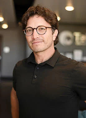Man with brown hair and glasses wearing a black polo shirt stands indoors with blurred lights and signage in the background.