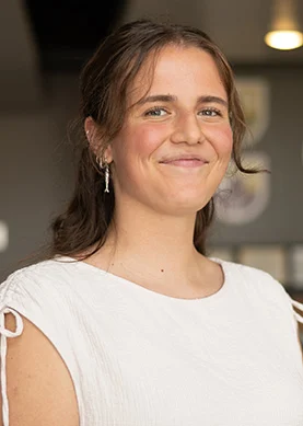 A woman with long brown hair wearing a white blouse and drop earrings stands indoors, smiling at the camera.