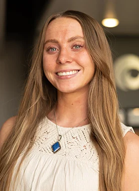 A woman with long, straight light brown hair wearing a sleeveless white top and a blue pendant necklace smiles at the camera indoors.
