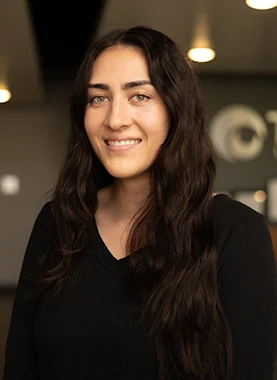 Woman with long dark hair and a black shirt smiles at the camera indoors with blurred lights in the background.