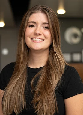 Young woman with long brown hair wearing a black shirt, smiling, standing indoors with blurred lights and wall in the background.