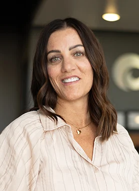 Woman with shoulder-length brown hair wearing a light-colored blouse and gold necklace, smiling indoors with blurred background.