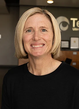 A woman with short blonde hair wearing a black top smiles at the camera in an office setting with certificates on the wall behind her.